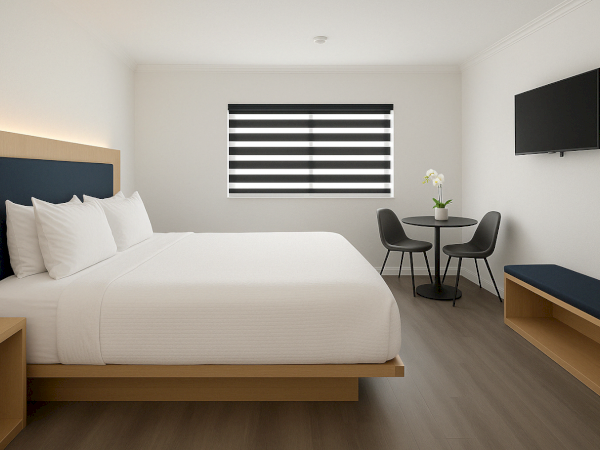 A modern hotel room with a large bed, black and white decor, a wall-mounted TV, small table with chairs, and a window with blinds.
