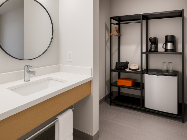 A hotel room corner with a sink, mirror, shelving unit, fridge, coffee maker, phone, and a hanging clothing rail.
