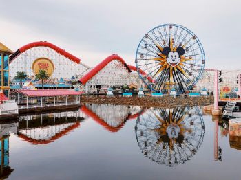 An amusement park scene features a ferris wheel and roller coaster reflected in calm water, under a cloudy sky.