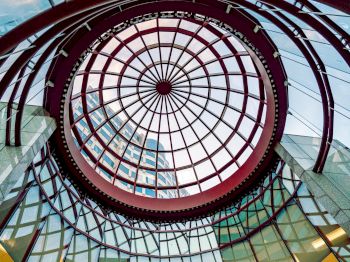 The image shows a glass dome with a geometric metal framework, viewed from below, with a building visible through the structure.