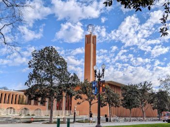 The image shows a campus scene with a brick building, a tall tower with a globe on top, trees, and a blue sky with clouds.