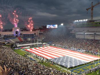 A large American flag on a football field, surrounded by a crowd and fireworks, creating a patriotic stadium atmosphere.