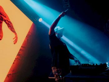 A DJ is performing on stage under blue lighting, with visuals on a screen behind them.
