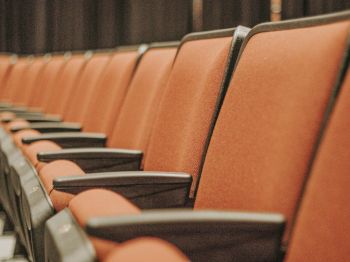 The image shows a row of empty, orange theater seats with black armrests, against a dark background.