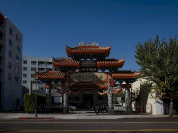 The image shows an ornate entrance gate with traditional Asian architectural elements, located between modern buildings against a clear blue sky.
