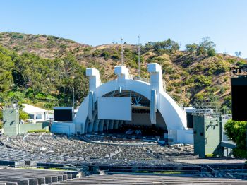 An open-air amphitheater with a large stage, surrounded by hills and empty seating rows, under a clear blue sky.