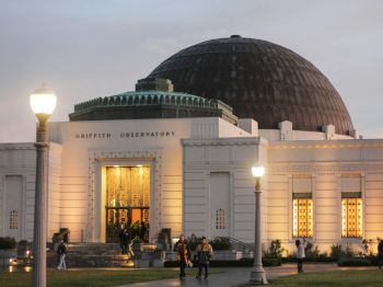 The image shows the Griffith Observatory with its dome, illuminated lamps, and people near the entrance.
