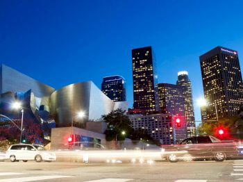 A vibrant cityscape at dusk with modern buildings, light trails from cars, and a distinctive metallic architecture on the left side.