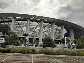The image shows the exterior of SoFi Stadium, with a curved roof and surrounding columns.