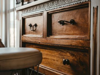The image shows a wooden dresser with decorative handles and a stool with a cushioned seat, in a warmly lit room.
