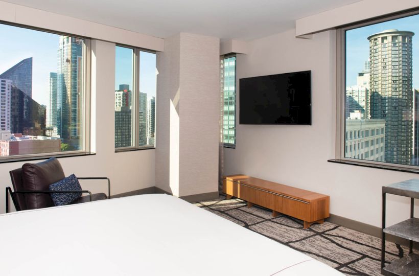 The image shows a modern room with large windows, a bed, a chair, a TV, and a view of city skyscrapers.