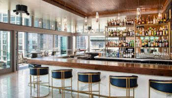 This image shows a modern bar with a sleek design, featuring high shelves filled with bottles, counter seating, and large windows with city views.