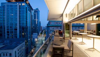 An outdoor balcony with tables and chairs overlooking a cityscape of high-rise buildings at dusk, featuring modern architecture and street lights.