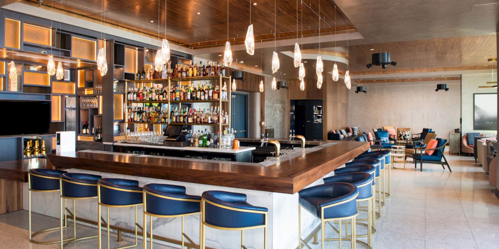 A modern bar with blue barstools, hanging lights, a well-stocked shelf, and a seating area in the background, featuring cozy chairs.