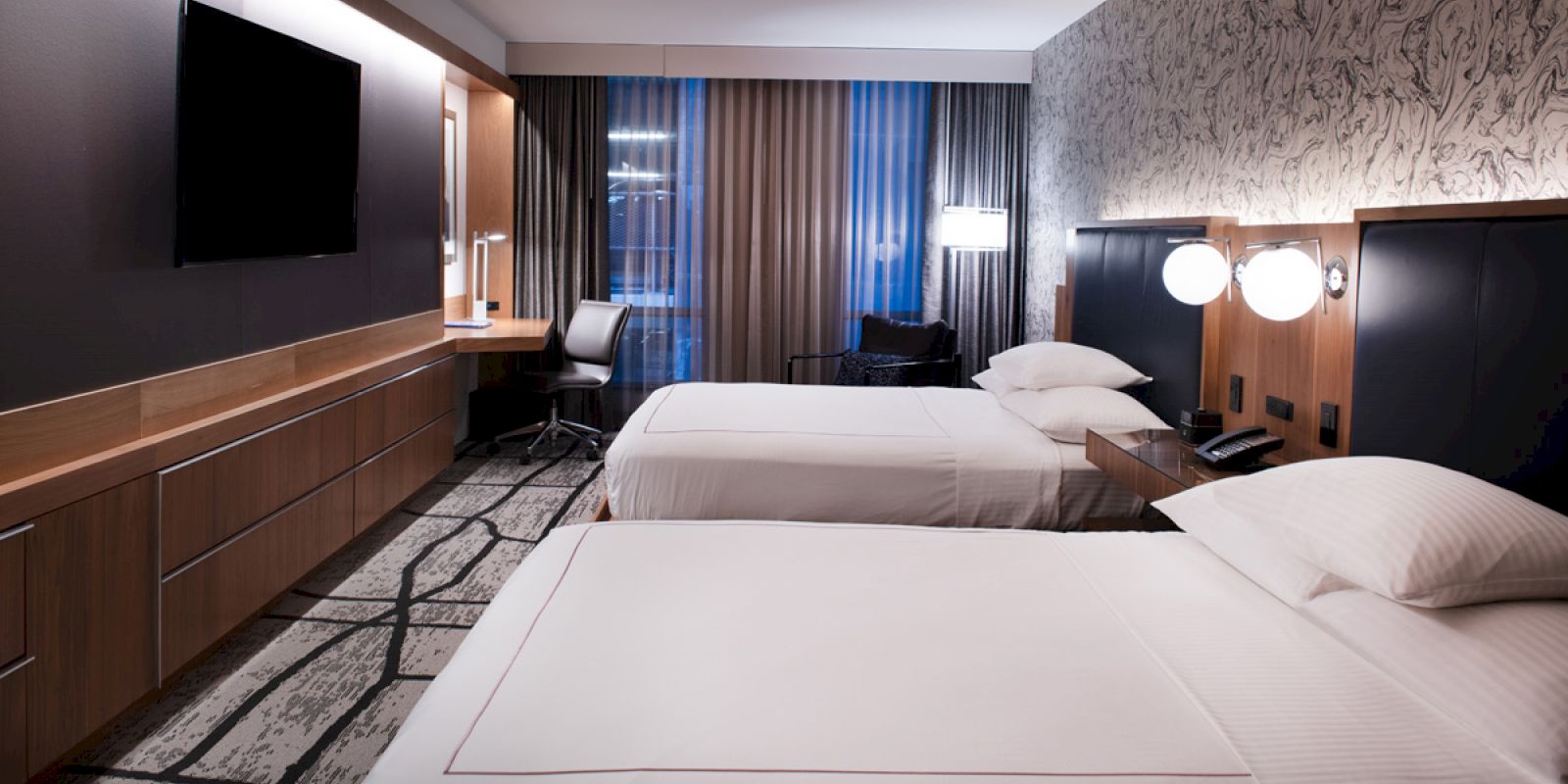A modern hotel room features two double beds, a wall-mounted TV, a desk with a chair, and large windows with curtains.