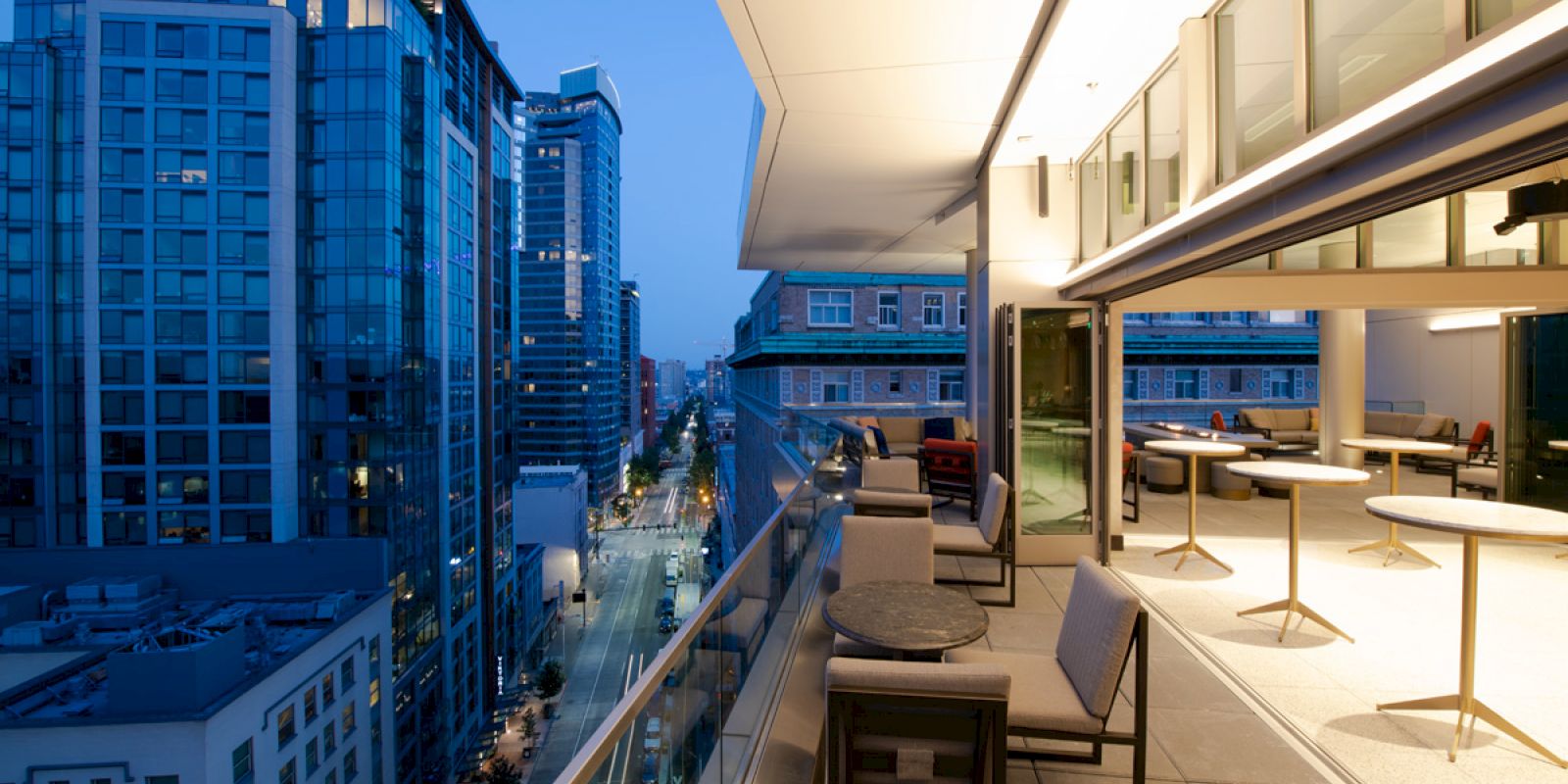 A modern cityscape at dusk from a high-rise balcony with tables, chairs, and an open indoor area, overlooking buildings and a street below.