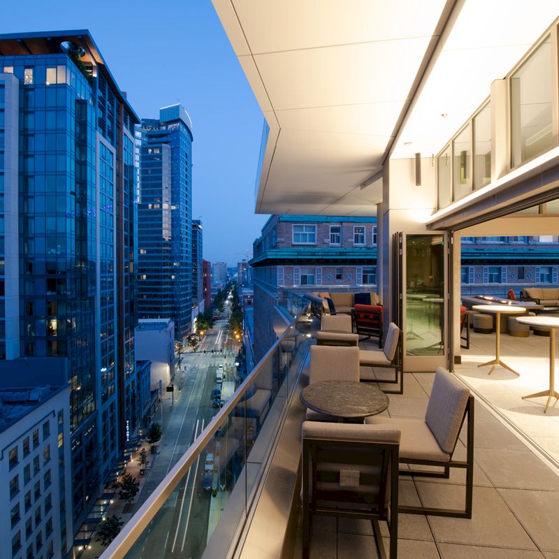 The image shows a modern cityscape at dusk, with a rooftop terrace featuring tables, chairs, and a glass railing overlooking high-rise buildings.