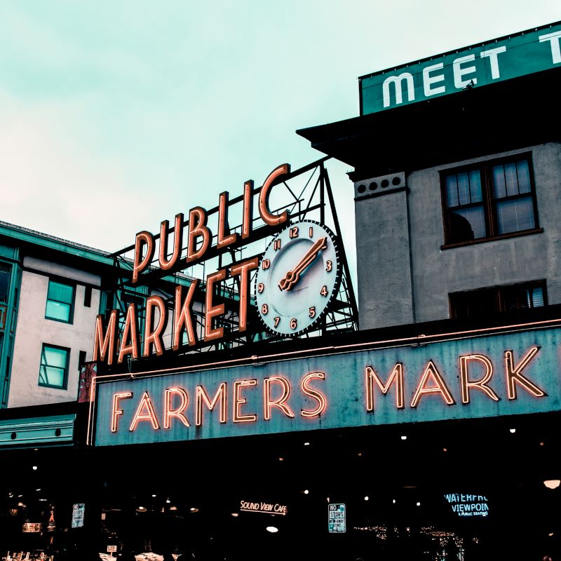 A neon sign reading "Public Market" with a clock, and another sign below it reading "Farmers Market," on buildings in a public market area.