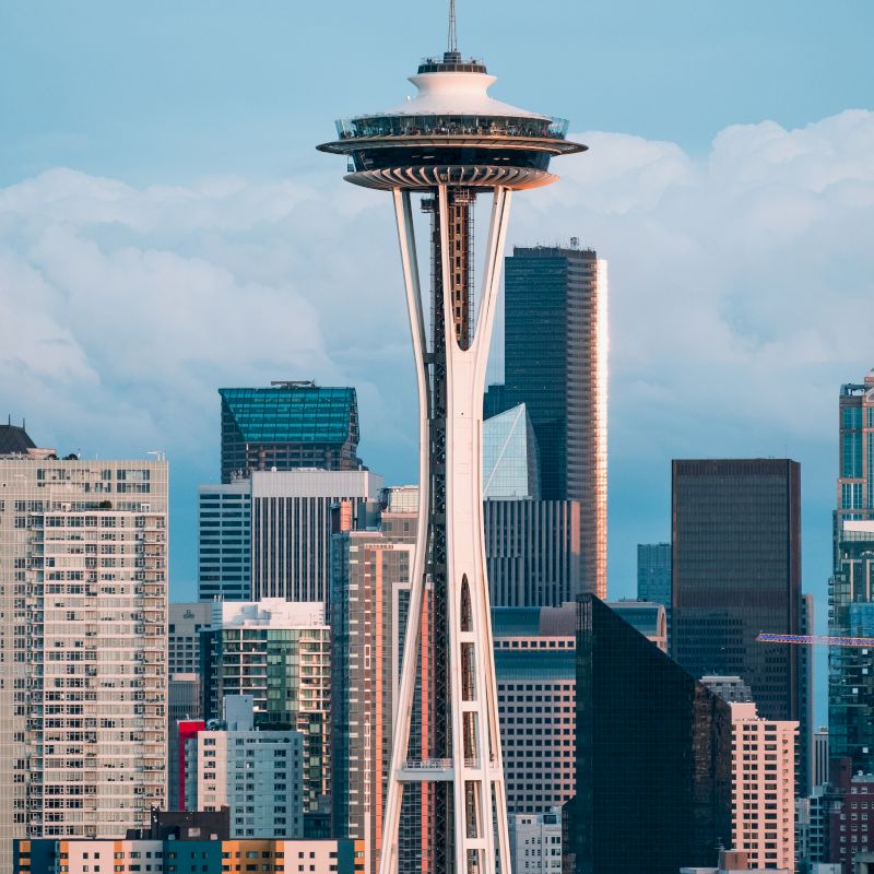 This image shows the Space Needle tower in Seattle, with surrounding modern buildings and a partly cloudy sky.