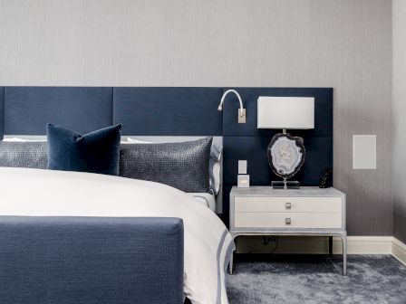 This image shows a modern bedroom with a blue and white color scheme, featuring a bed with a blue headboard, pillows, and a nightstand with a lamp and decor.