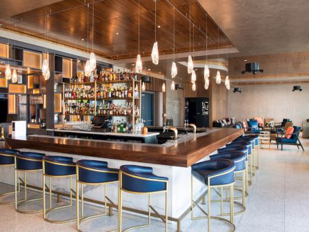 A modern bar with stylish bar stools, a fully stocked liquor shelf, pendant lights, and a lounge area with additional seating in the background.