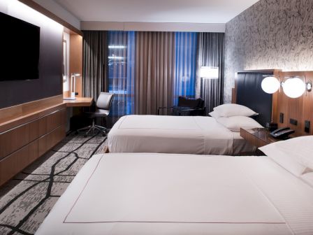 A modern hotel room with two beds, a desk, a large TV on the wall, and stylish decor consisting of a patterned wall and contemporary furnishings.