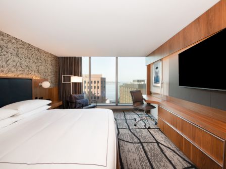 A modern hotel room with a large bed, an expansive window view of city buildings, a wall-mounted TV, and a desk with a chair near the window.
