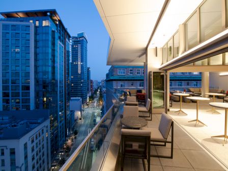 A modern, high-rise balcony at dusk overlooking a city street, furnished with tables and chairs. The adjacent interior is well-lit and stylish.