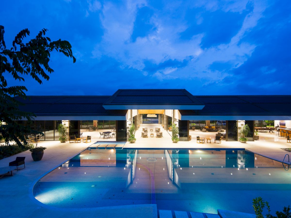 A luxurious poolside area at dusk, surrounded by a modern building with open, inviting spaces and outdoor seating.