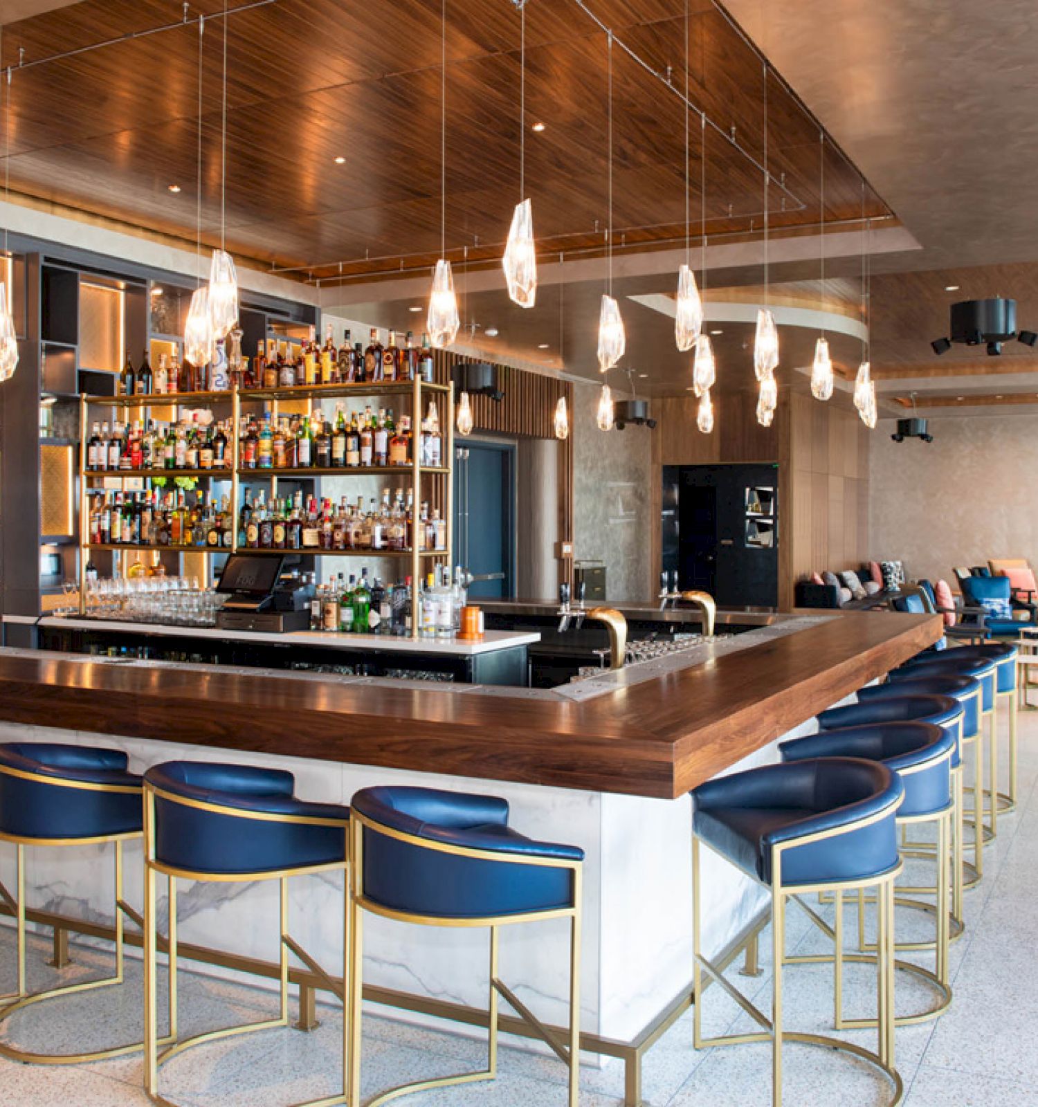 This image shows a modern bar with a sleek design: blue barstools with gold legs, hanging lights, and a variety of bottles behind the counter.