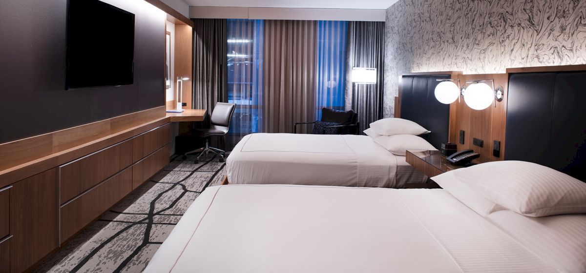 The image shows a modern hotel room with two beds, a flat-screen TV, a desk and chair, nightstands, and stylish wallpaper.