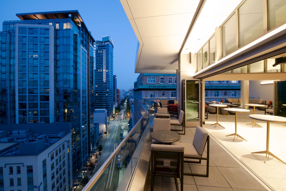 A modern outdoor balcony with tables and chairs overlooks a bustling cityscape at dusk, with tall buildings and a lit street below.