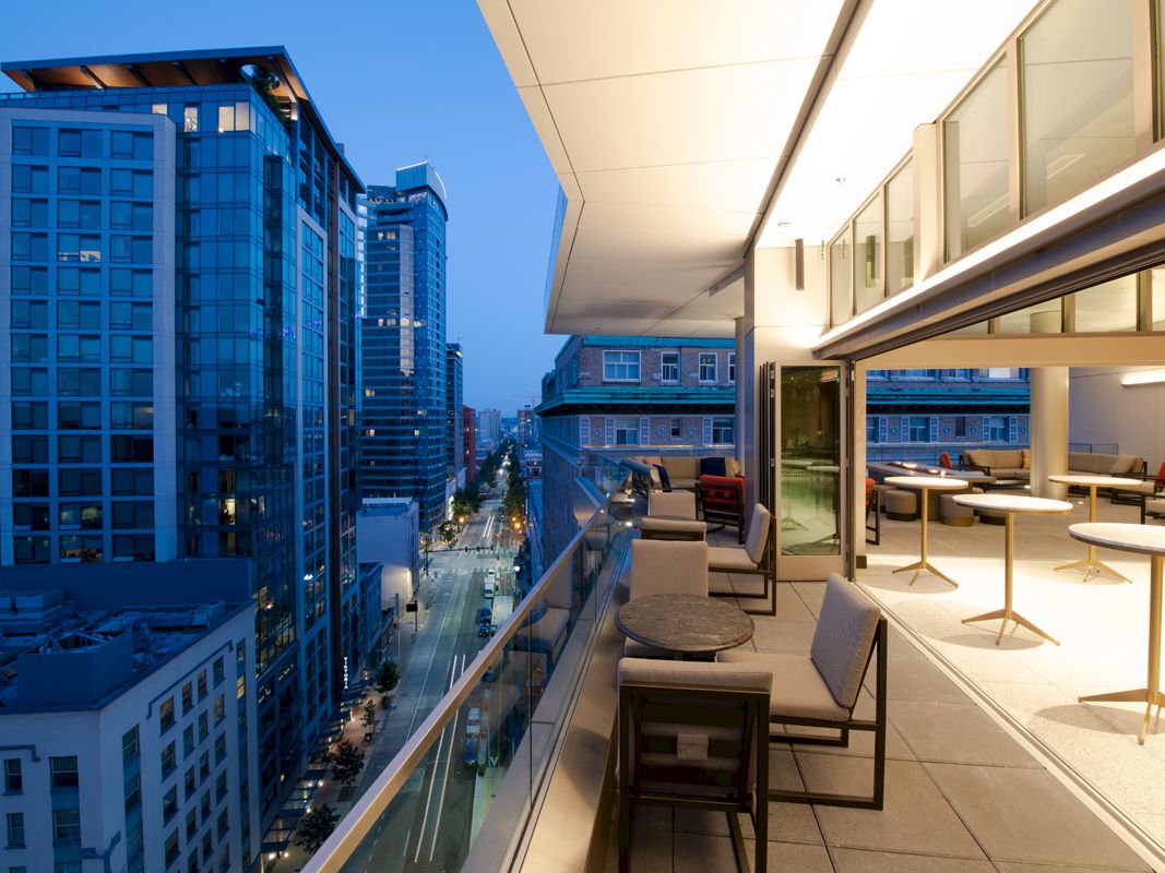 A modern outdoor balcony with tables and chairs overlooks a bustling cityscape at dusk, with tall buildings and a lit street below.