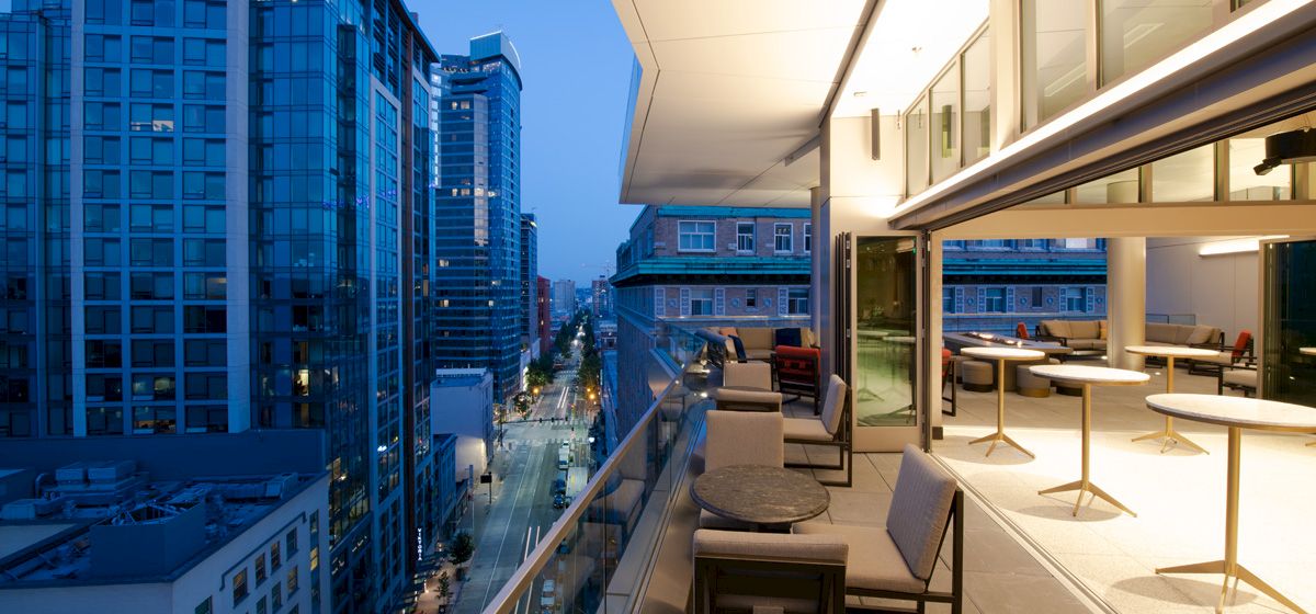 A modern outdoor balcony with tables and chairs overlooks a bustling cityscape at dusk, with tall buildings and a lit street below.