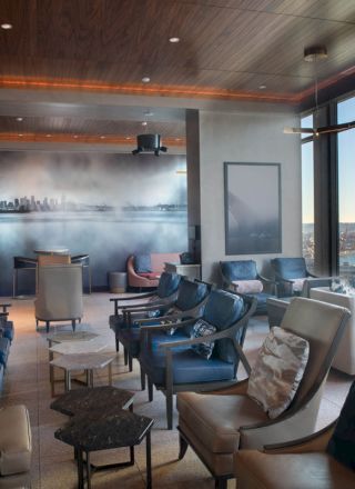A modern lounge with ample seating, large windows offering a coastal view, and a mural depicting a city skyline on the wall.