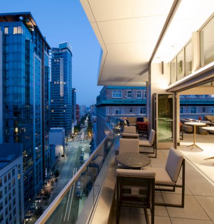 A modern balcony of a high-rise building with tables and chairs overlooks a city street with tall buildings during the evening.