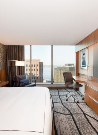 Modern hotel room with a large bed, wall-mounted TV, desk, chair, floor-to-ceiling window with a city view, and patterned walls and carpet.