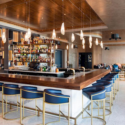 This image shows a modern bar with blue bar stools, a counter with various bottles, hanging lights, and seating areas in the background.
