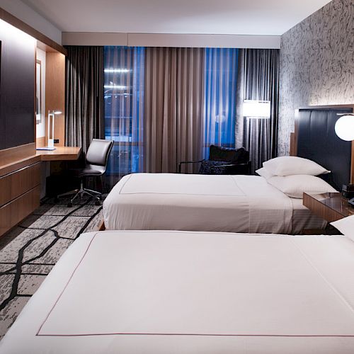 A modern hotel room with two beds, a wall-mounted TV, a desk with a chair, and lamps, featuring contemporary decor and large windows with curtains.