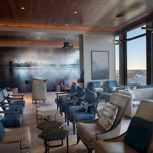 A modern lounge with comfortable seating, large windows with a view of the waterfront, and a mural of a city skyline on the back wall.
