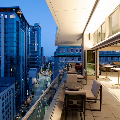A modern cityscape viewed from a high-rise balcony at dusk, featuring tall buildings, bright lights, and a chic outdoor seating area.