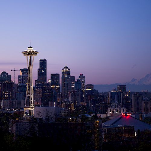 The image showcases the Seattle skyline at dusk, featuring the Space Needle prominently with Mount Rainier visible in the background, against a fading blue sky.