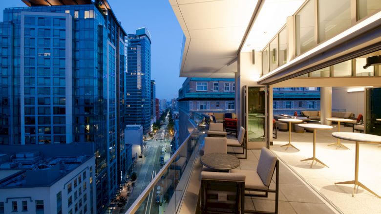 A modern rooftop with tables, chairs, and an open indoor area overlooks a city street lined with tall buildings, illuminated at dusk.