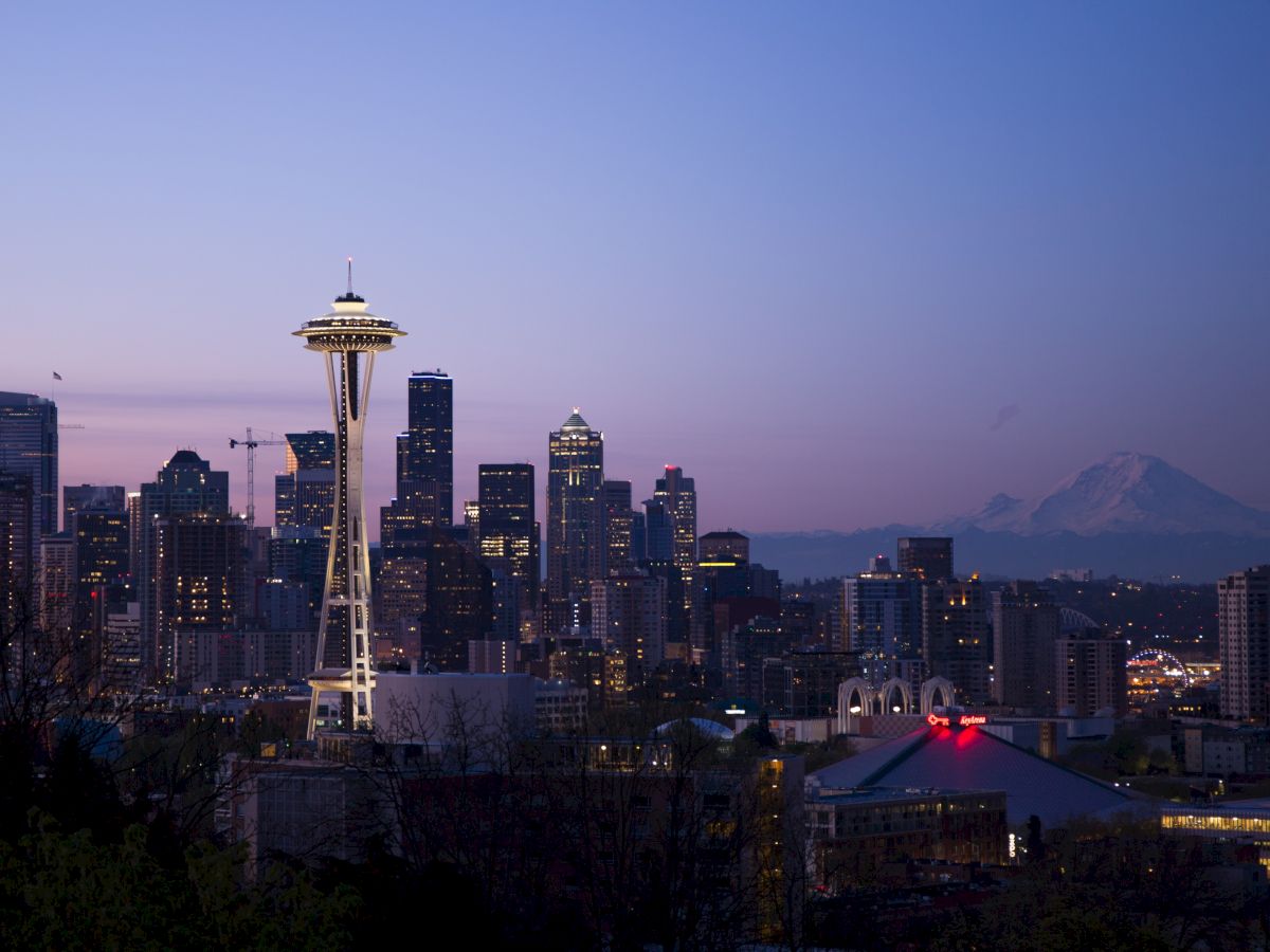 A view of Seattle's skyline with the Space Needle illuminated at dusk, against a backdrop of mountain peaks in the distance, under a fading blue sky.