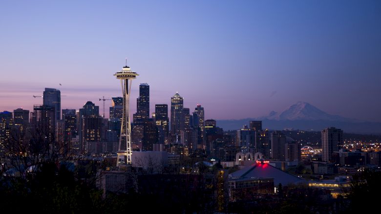 A view of Seattle's skyline with the Space Needle illuminated at dusk, against a backdrop of mountain peaks in the distance, under a fading blue sky.