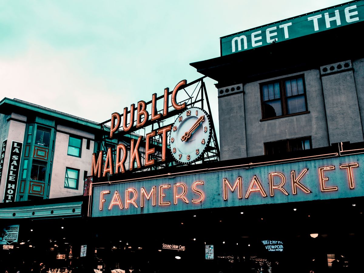 The image shows the entrance to a farmers market with neon signs reading "PUBLIC MARKET" and "FARMERS MARKET," located in an urban area.