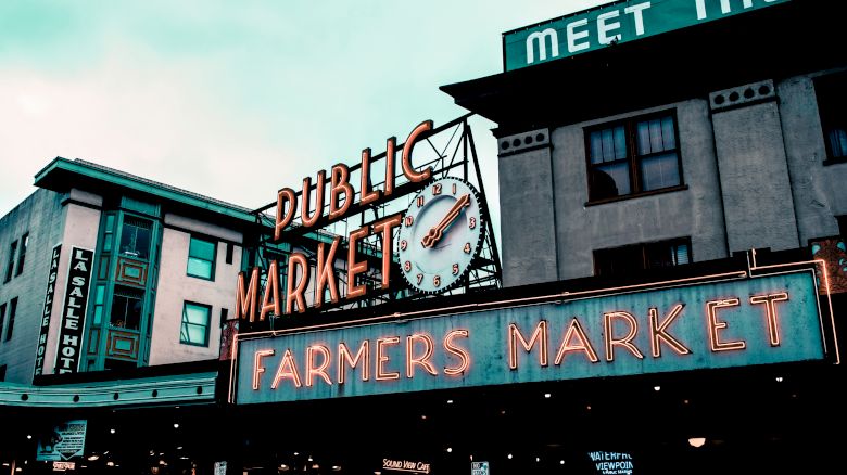 The image shows the entrance to a farmers market with neon signs reading "PUBLIC MARKET" and "FARMERS MARKET," located in an urban area.