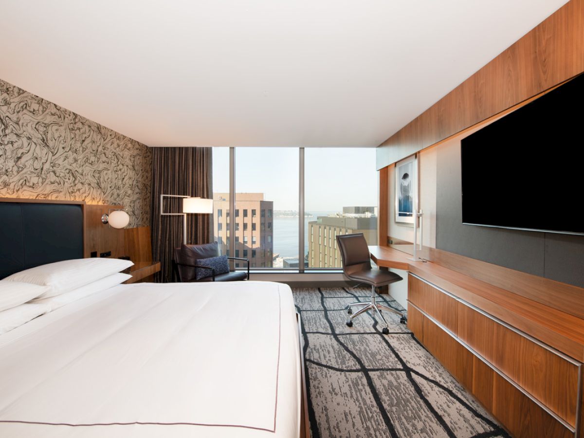 Modern hotel room with a large bed, desk, chair, flat-screen TV, and large window with a city view.