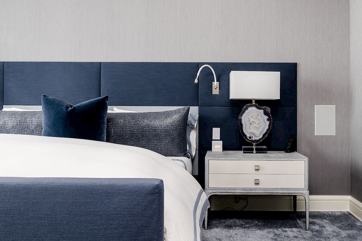 This image shows a modern bedroom with a blue upholstered headboard, white and gray bedding, and a nightstand with a lamp and decorative items.