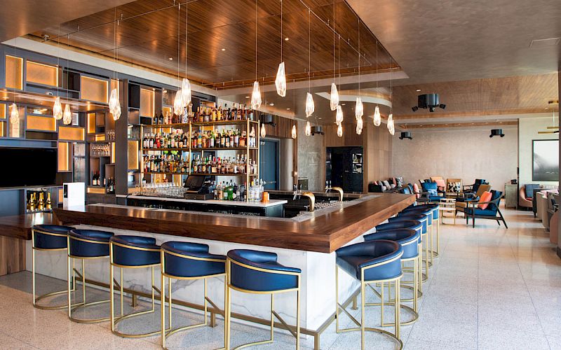 A modern bar with blue stools, a well-stocked assortment of drinks, stylish hanging light fixtures, and a cozy seating area in the background.